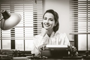 35506381 - 1950s style secretary working at office desk and smiling with hand on chin