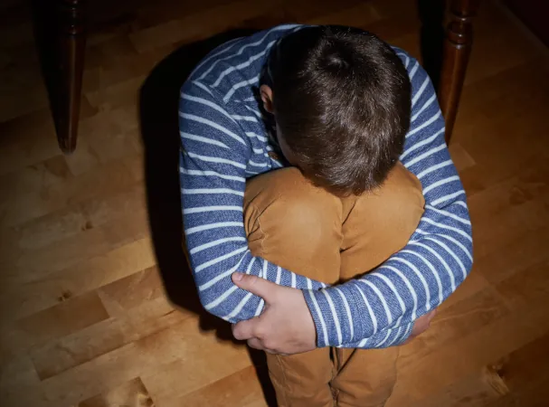 An upset boy sitting on floor with his knees drawn up, arms wrapped around his knees and face buried in his knees.