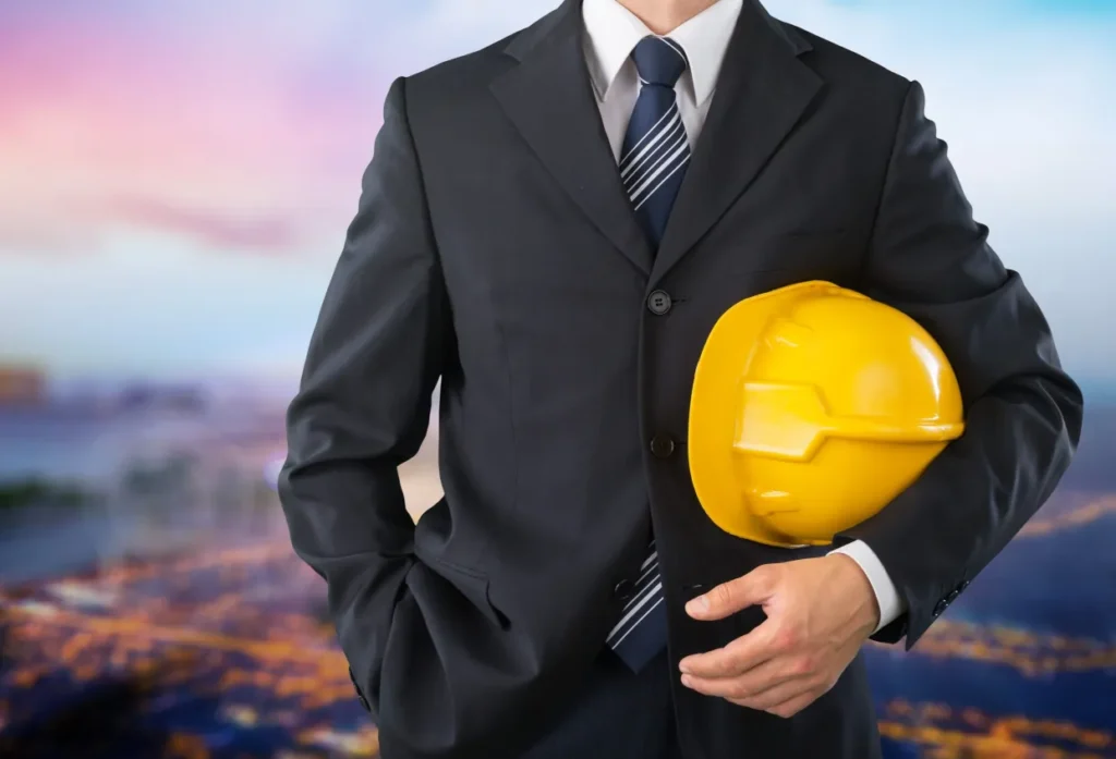 A confident professional in a suit holds a yellow hard hat, symbolizing the blend of business acumen and construction expertise as the city skyline reflects the hues of dusk behind him.